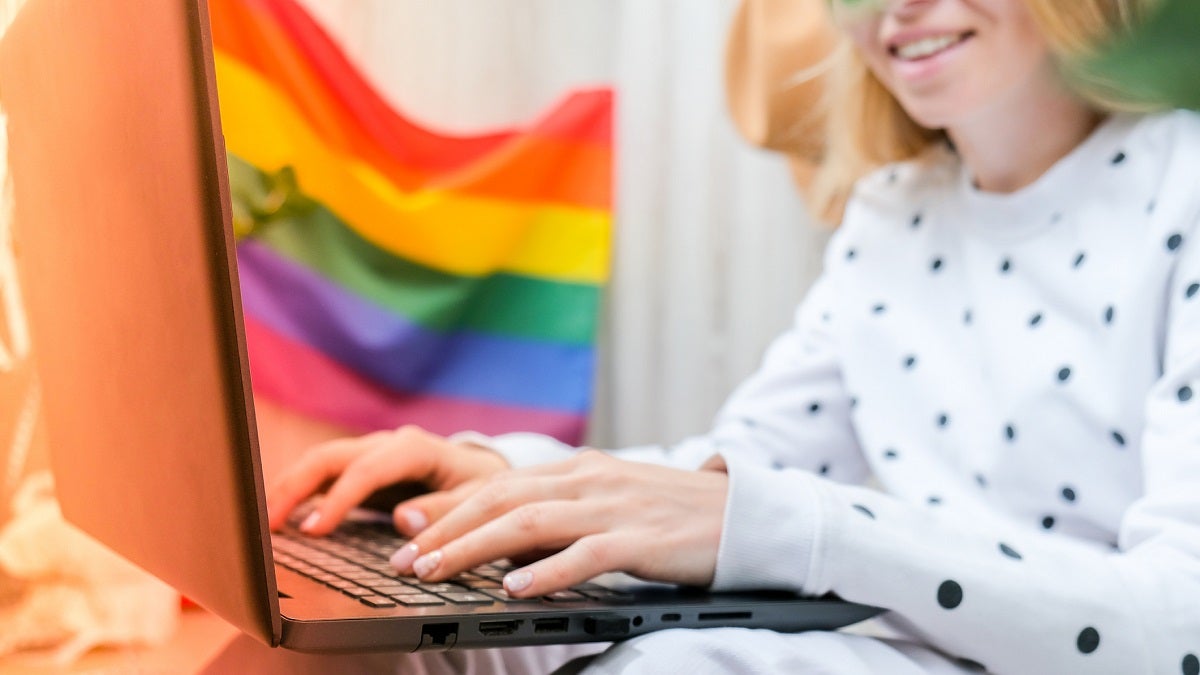 Young Millennial Hippie Woman Sitting On Balcony Using Laptop. Lgbtq Rainbow Flag On Background.