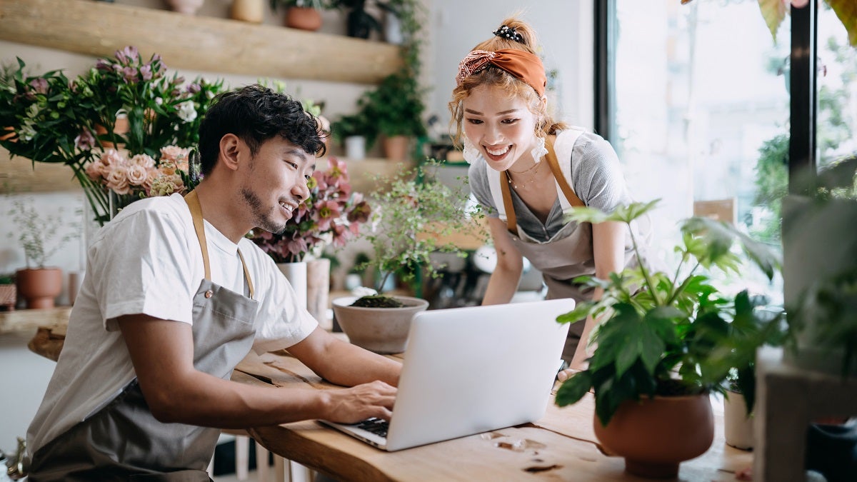 Smiling young Asian couple, the owners of small business flower shop, discussing over laptop on counter against flowers and plants. Start-up business, business partnership and teamwork. Working together for successful business